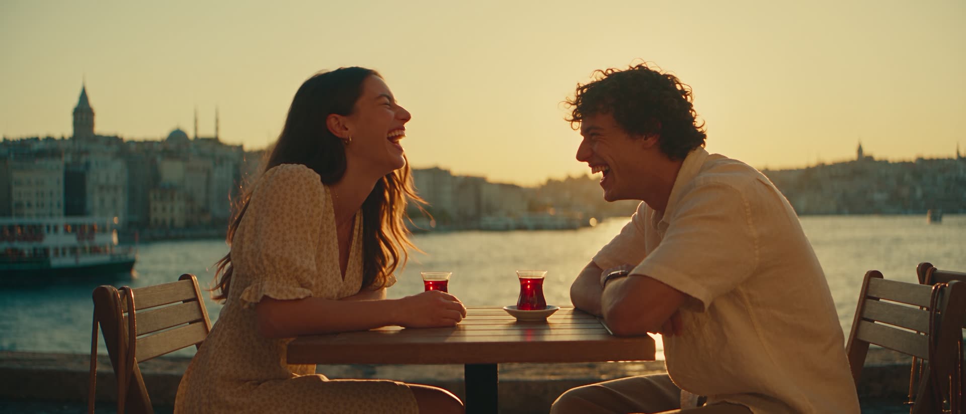 Zwei Menschen lachen bei Sonnenuntergang an einem Tisch am Bosporus in Istanbul, vor ihnen zwei Gläser türkischer Tee